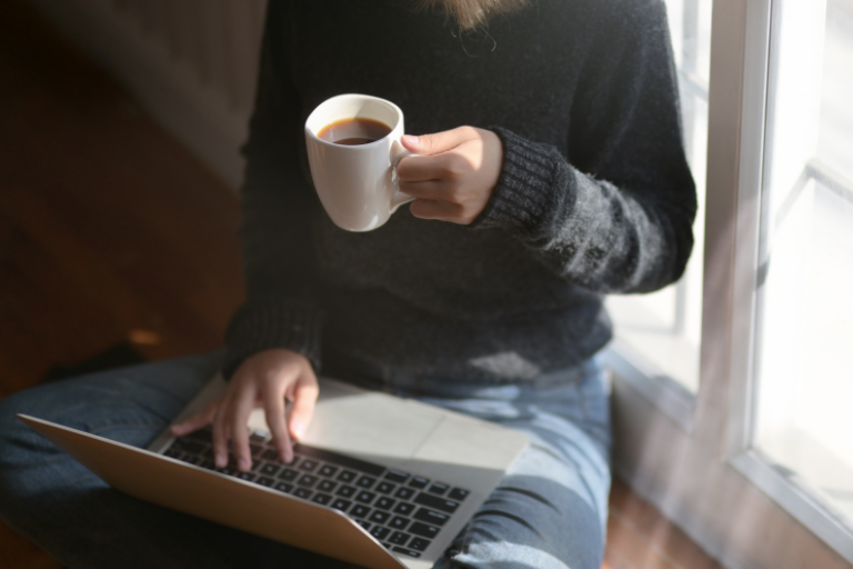 窓辺に胡座をかいてコーヒーを飲みながらノートパソコンを操作している女性